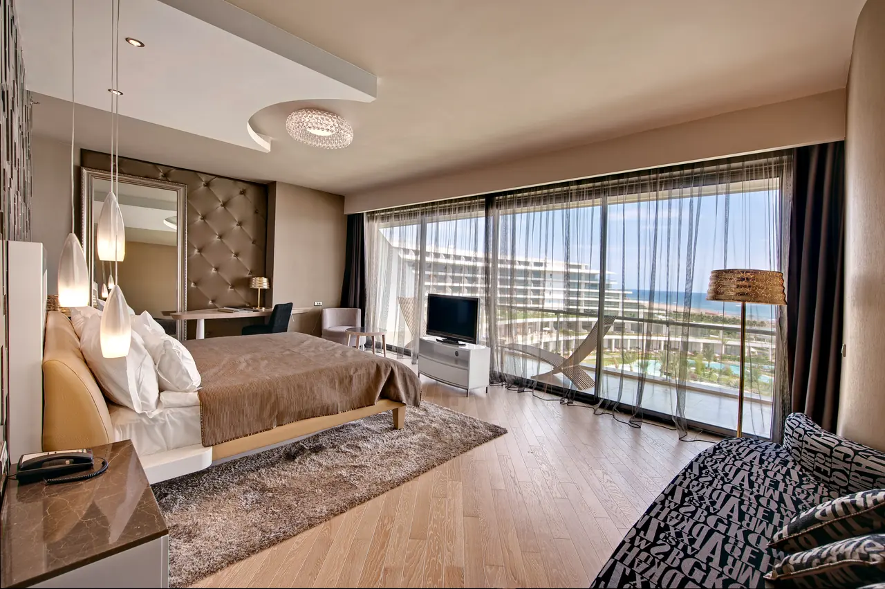 Maxx Royal Belek — Bedroom with a glass wall overlooking the sea and resort