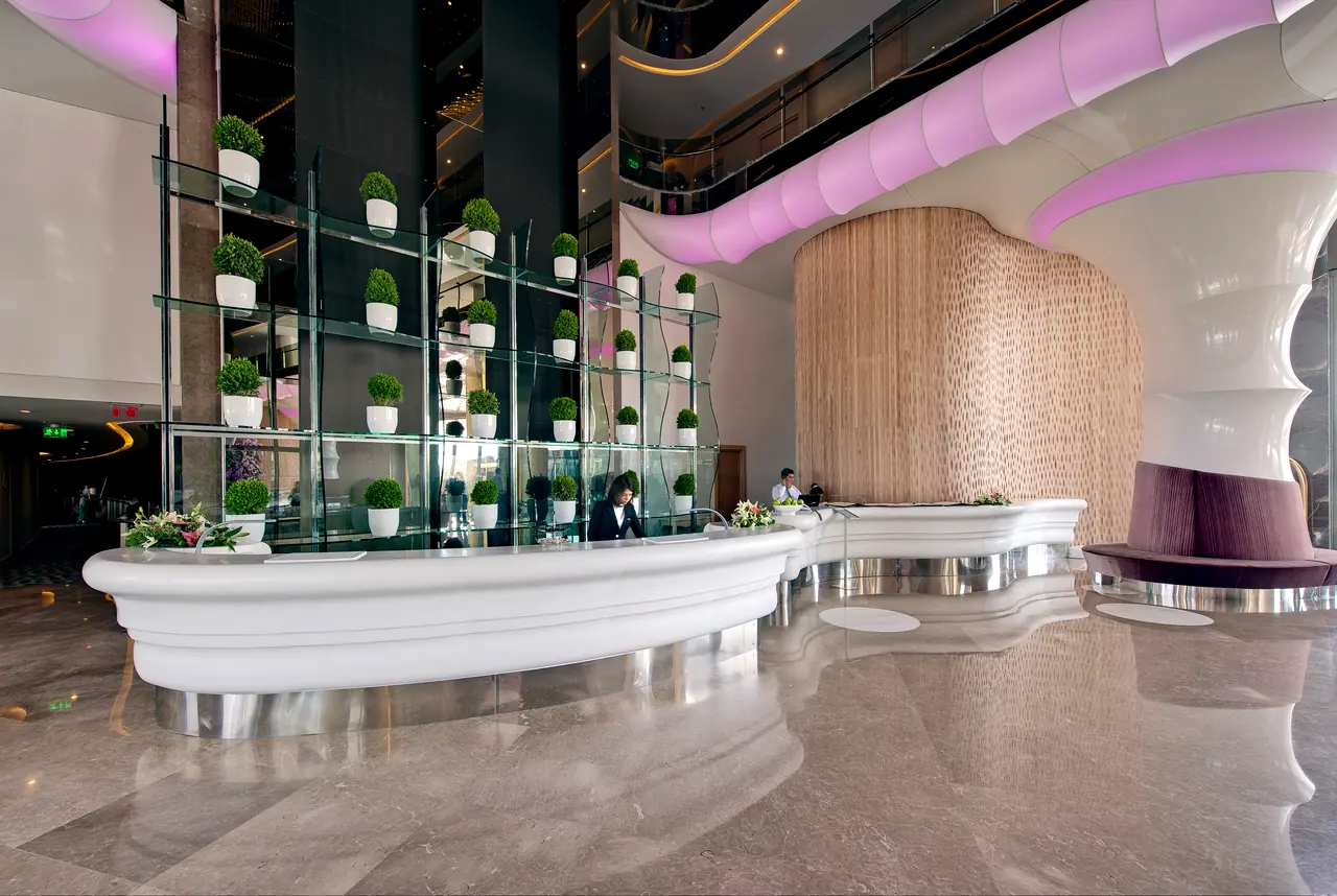 Maxx Royal Belek — Reception with a curved white desk and planted shelving